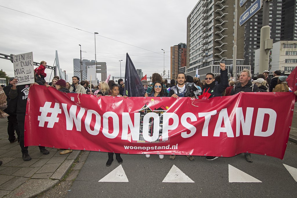 Demonstranten lopen met grote banner met #woonopstand door Rotterdam. De Erasmusbrug is zichtbaar op de achtergrond.